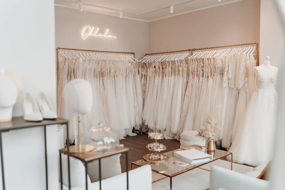 Willkommensbereich der maleika Brautboutique Krefeld mit Ohlala-Leuchtschriftzug, Brautkleidern an goldenen Kleiderstangen und Sitzecke mit Accessoires